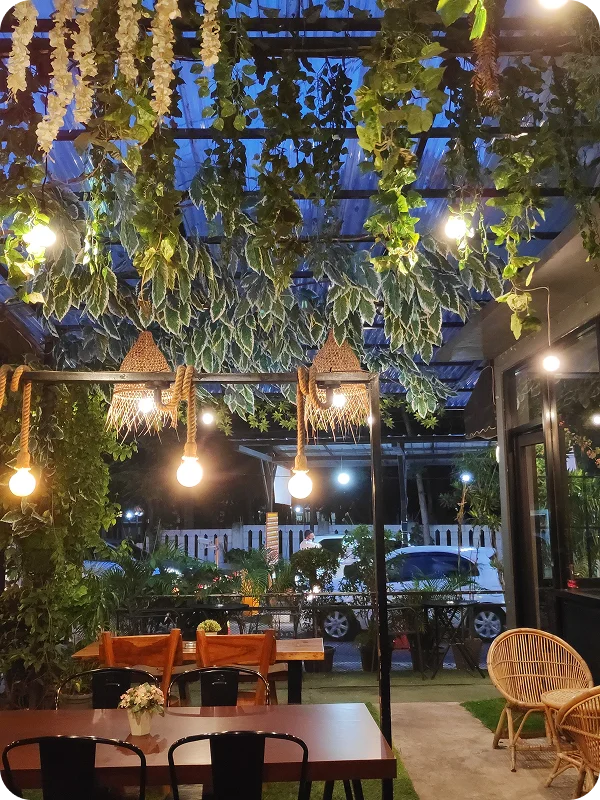 Een weelderig restaurantterras bij schemering, vol met hangplanten, warme lichtslingers, houten tafels, zwarte stoelen en gezellige rieten zitjes.